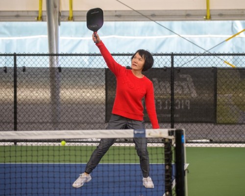 A woman in a red sweater prepares to hit a pickleball with a paddle on an indoor court.