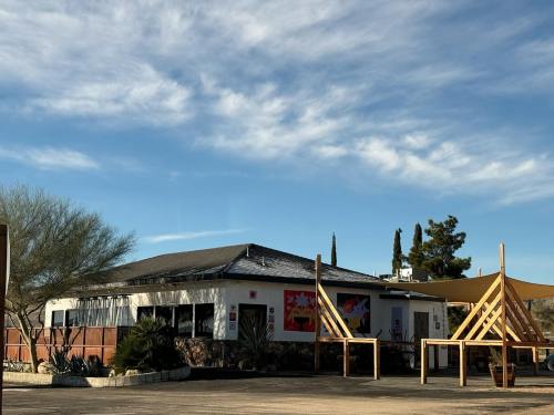 A modern building with a flat roof, surrounded by desert plants and wooden structures, under a clear blue sky.