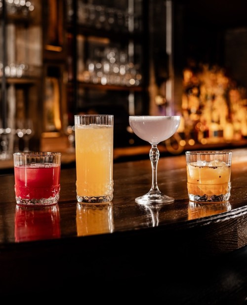 Four colorful cocktails on a wooden bar, featuring a red drink, a yellow drink, a clear cocktail, and an orange drink.