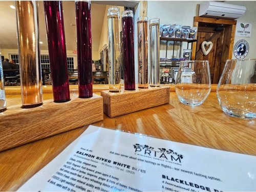 A wooden tasting flight holder with colorful wine samples, two glasses, and a menu on a table in a cozy setting.