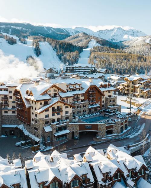 A snowy mountain resort with cozy buildings, surrounded by pine trees and slopes under a clear blue sky.