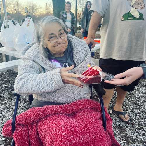 An elderly woman in a cozy sweater holds a slice of cake with a candle, smiling while seated in a wheelchair.