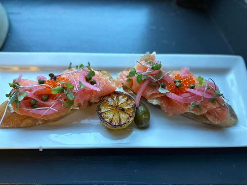 Two pieces of toast topped with smoked salmon, capers, pickled onions, and microgreens, served with a grilled lemon slice.