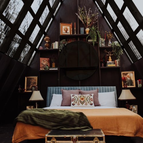 Cozy bedroom with a large bed, decorative pillows, plants, and a stylish bookshelf in a modern, triangular space.