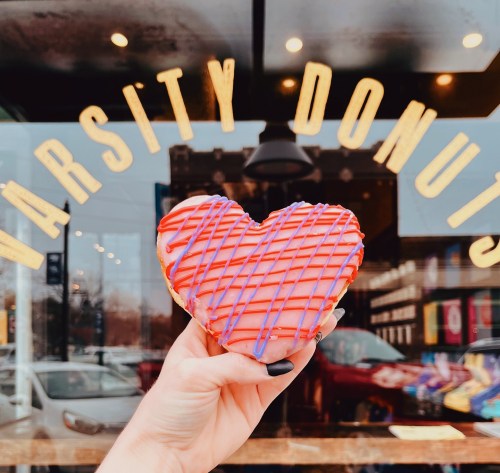 A hand holds a heart-shaped donut with red and blue icing in front of a shop window that reads "Varsity Donuts."