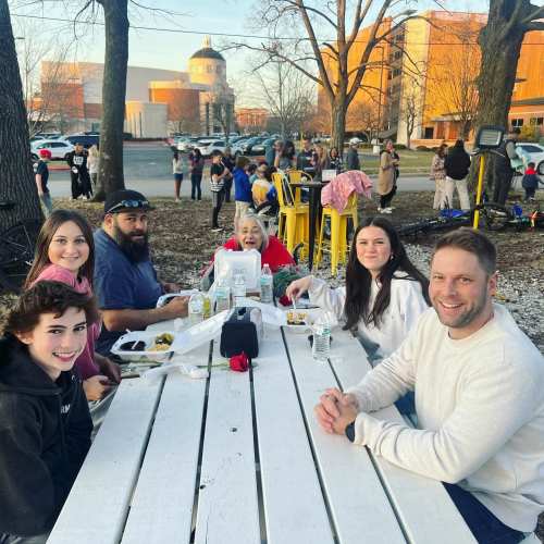 A group of six people enjoying a meal outdoors at a picnic table, with trees and a crowd in the background.