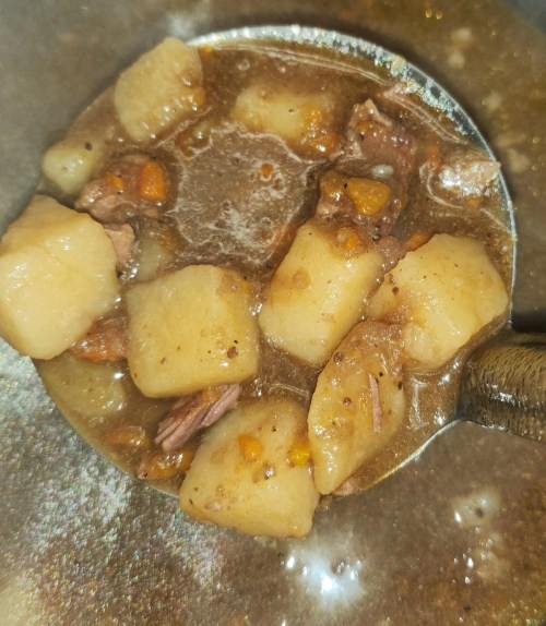A close-up of a ladle filled with hearty beef stew, featuring chunks of meat, potatoes, and carrots in a rich broth.