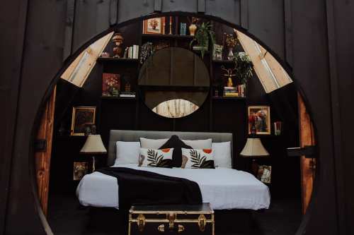 Cozy bedroom with a round window, featuring a bed, decorative pillows, bookshelves, and warm lighting.