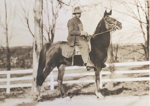 A man in period clothing sits on a horse, posed beside a white fence with bare trees in the background.
