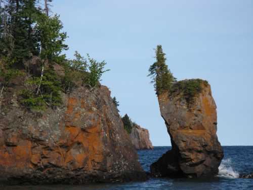 A tall rock formation with a small tree on top, surrounded by water and rocky shores under a clear blue sky.