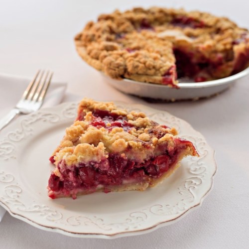 A slice of cherry pie with a crumbly topping on a decorative plate, next to a fork and the pie in the background.