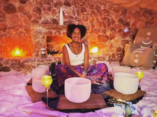 A woman sits in a cozy, salt cave surrounded by crystal singing bowls, with warm lights and a stone fireplace in the background.
