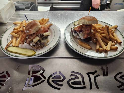 Two plates with sliders and fries, one topped with bacon and cheese, the other with pulled pork, on a metal counter.