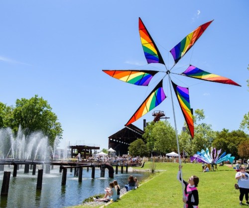 A colorful rainbow wind spinner stands by a pond, with people enjoying a festival in the background under a clear blue sky.