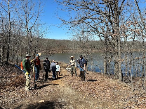 A group of people walking along a trail by a lake, surrounded by bare trees and a clear blue sky.