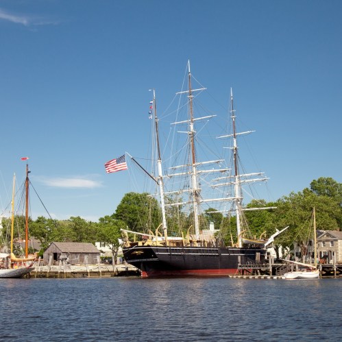 A tall ship with multiple masts docked by the shore, surrounded by smaller boats and greenery under a clear blue sky.