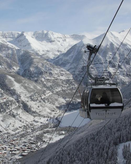 A gondola ascends over a snowy mountain landscape, with a valley and town visible below.