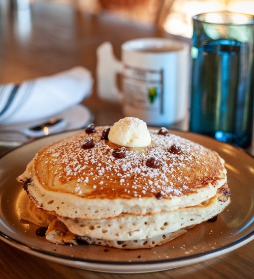 A stack of fluffy pancakes topped with butter and chocolate chips, dusted with powdered sugar, on a plate.