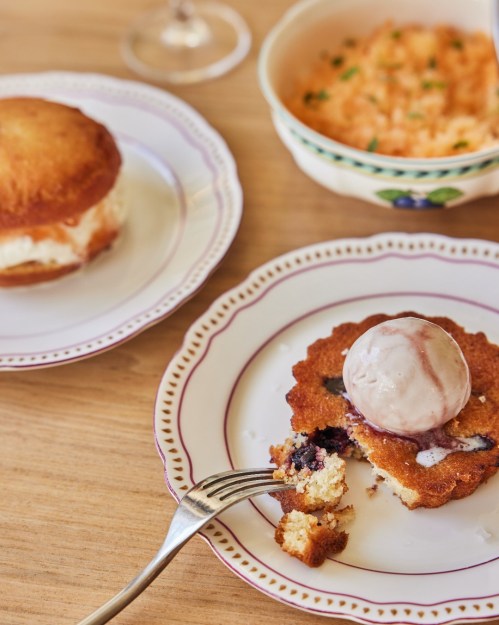 A dessert plate featuring a blueberry cake topped with ice cream, alongside a bun and a bowl of creamy dessert.