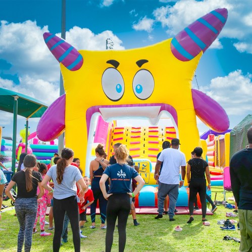 A colorful inflatable slide with a smiling monster face, surrounded by children and adults enjoying a sunny day.