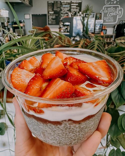 A hand holding a clear cup of layered dessert with chia seeds, yogurt, and fresh strawberries on top.