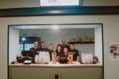 A group of six staff members smiling behind a bar, with drinks and a cash register visible.