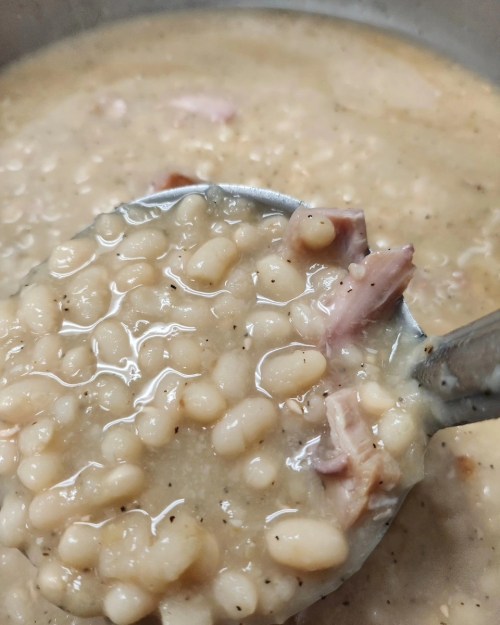 A close-up of a ladle filled with creamy white bean soup, featuring pieces of ham and a smooth broth.