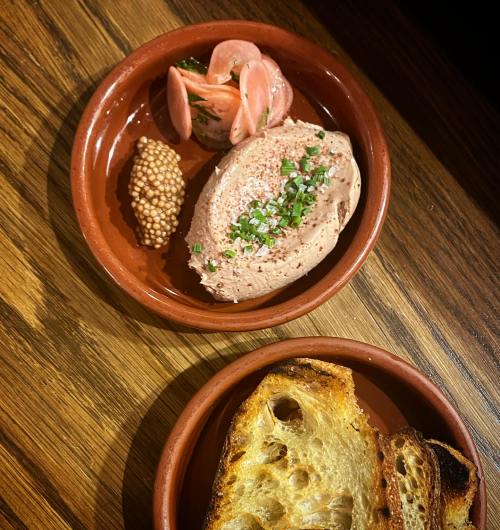 A clay dish with pâté, pickled vegetables, mustard, and toasted bread on a wooden table.