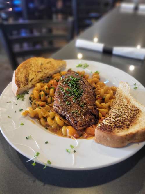 A plate of pasta with a seasoned fish fillet, garnished with herbs, and served with two slices of toasted bread.