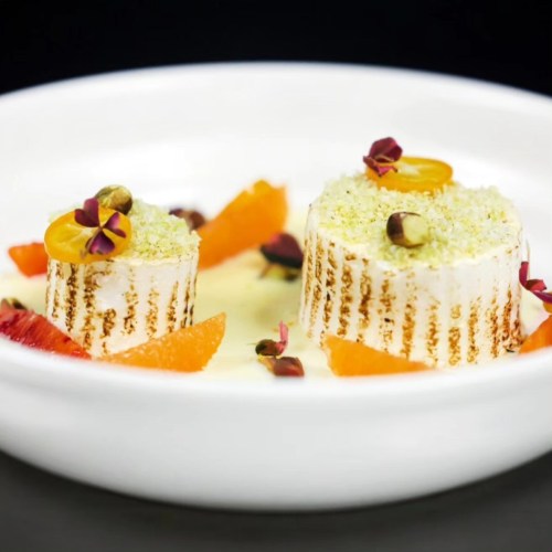 A dessert featuring two cylindrical cakes topped with pistachios and edible flowers, surrounded by citrus segments.