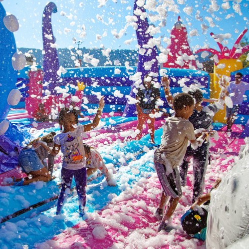 Children playing in a colorful foam-filled area, enjoying a fun outdoor activity on a sunny day.