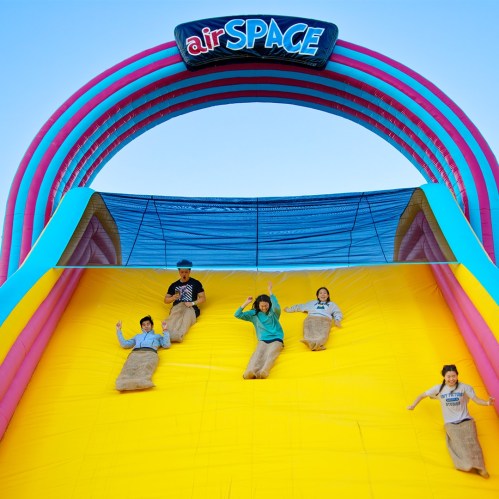 A colorful inflatable slide with people in burlap sacks sliding down against a clear blue sky.