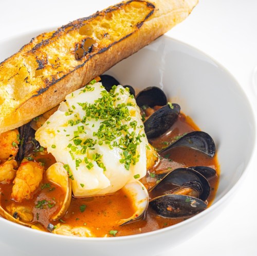 A bowl of seafood stew with shrimp, clams, mussels, and a piece of white fish, topped with herbs and served with toasted bread.