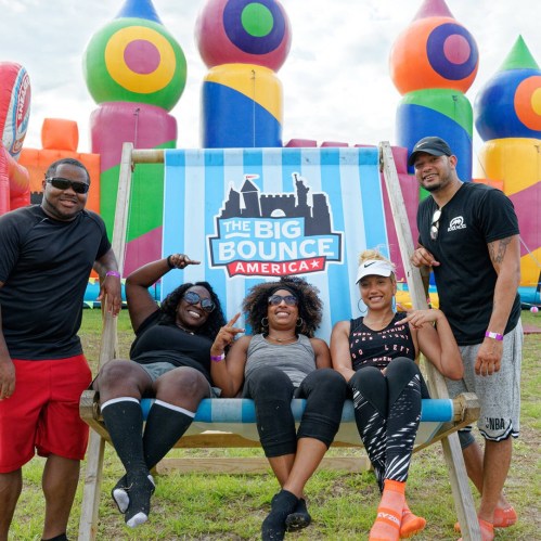 A group of five friends poses on a large swing in front of colorful inflatable structures at a bounce event.