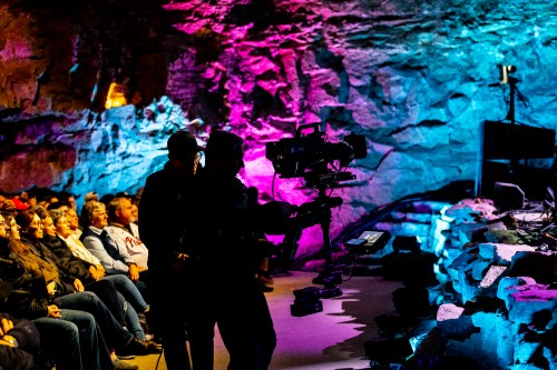 A camera operator films an audience in a colorful cave setting, with vibrant lights illuminating the rock walls.