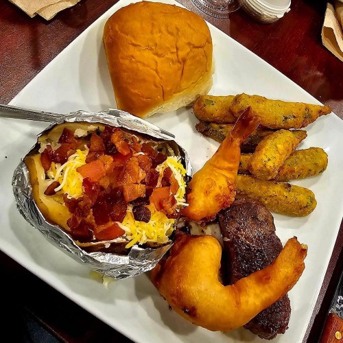 A plate featuring a baked potato with toppings, shrimp, steak, fried plantains, and a dinner roll.