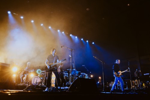 A band performs on stage with dramatic lighting and smoke, featuring guitarists and a drummer.