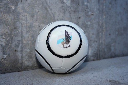 A white soccer ball with a black design and the Minnesota United FC logo against a concrete background.