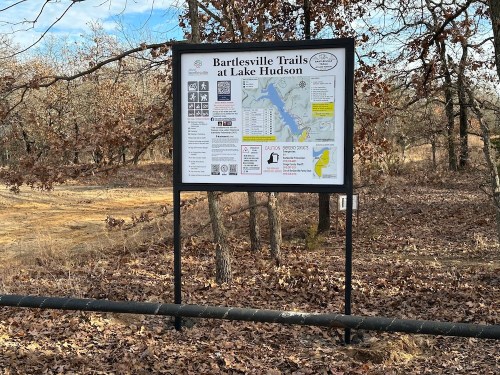 Sign for Bartlesville Trails at Lake Hudson, featuring maps and trail information, surrounded by bare trees and fallen leaves.