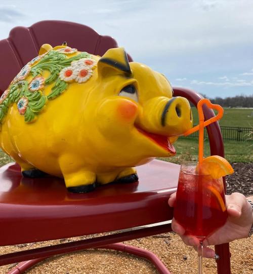 A cheerful yellow pig statue sits on a red chair next to a hand holding a colorful drink with a heart-shaped straw.