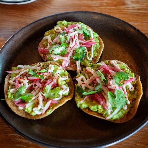 Three tostadas topped with guacamole, pickled onions, cilantro, and drizzled with cream on a dark plate.