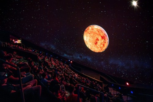 A planetarium audience watches a large, glowing planet projected on a starry dome ceiling.