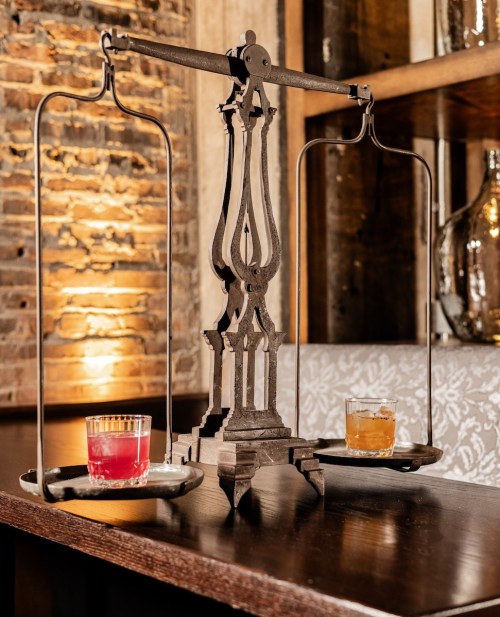 A vintage scale balances two glasses of colorful drinks on a wooden table against a rustic brick wall.