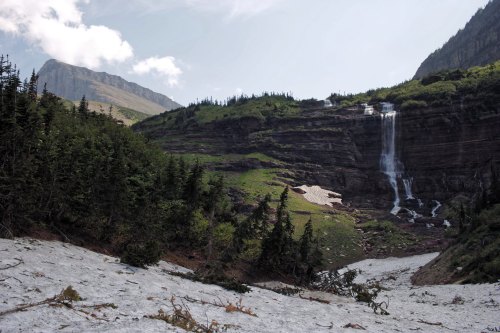 A scenic view of a waterfall cascading down rocky cliffs, surrounded by lush greenery and snow-covered ground.