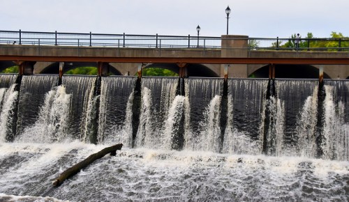 A waterfall cascades over a dam, with a bridge and lampposts in the background under a cloudy sky.