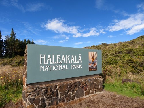 Sign for Haleakalā National Park against a blue sky with clouds and greenery in the background.