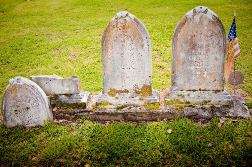 Three weathered gravestones in a grassy cemetery, with a small American flag beside one stone.