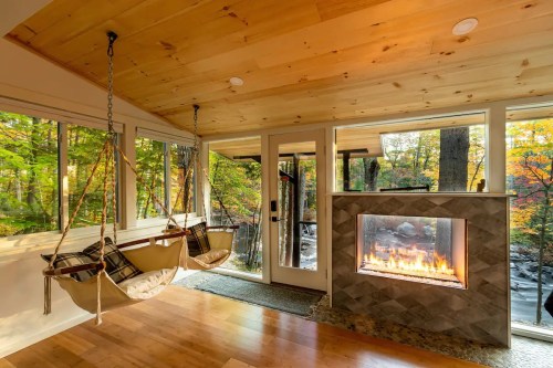 Cozy interior with hanging chairs, a stone fireplace, and large windows showcasing a vibrant forest view.