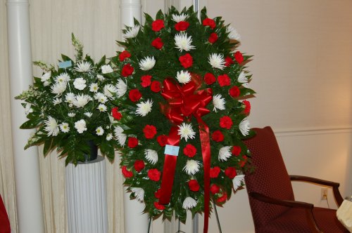 A floral wreath featuring red roses, white chrysanthemums, and greenery, adorned with a red ribbon.