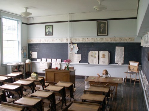 A vintage classroom with wooden desks, a chalkboard, and a child sitting at a desk, surrounded by educational materials.